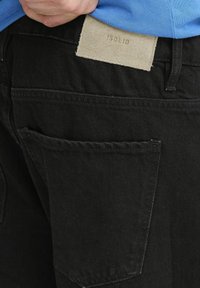 Człowiek podnosi niebieską koszulę włożoną w czarne dżinsy z beżową, prostokątną metką na pasku.