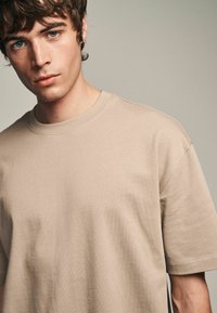 Beige Baumwoll-T-Shirt mit kurzen Ärmeln, Rundhalsausschnitt und einer weichen, glatten Textur. Merkmale ein minimalistisches Design und subtile Nähdetails.