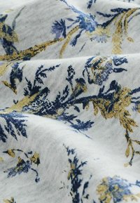 Tissu floral avec un fond blanc, présentant des motifs botaniques bleus et dorés. Texture douce avec des détails de couture visibles.