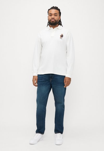 Λευκό πόλο μπλουζάκι με μακριά μανίκια, γιακά και κεντητό λογότυπο αρκούδας, σε συνδυασμό με σκούρο μπλε τζιν παντελόνι και λευκά αθλητικά παπούτσια.