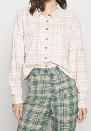 Kvinde iført beige og hvid ternet skjorte med knapper, let stoppet ind i grønne og beige ternede bukser.
