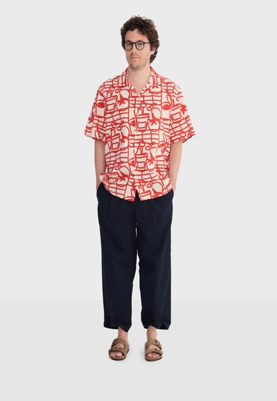 Homme aux cheveux bouclés et lunettes portant une chemise à manches courtes rouge et blanche à motifs, un pantalon ample foncé et des sandales marron, debout les mains dans les poches.