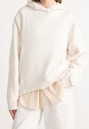 Personne portant un sweat à capuche beige clair superposé, une chemise beige rayée en dessous et un pantalon blanc cassé, avec une main dans la poche.