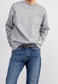 Sudadera gris de algodón, con cuello redondo y un pequeño logo. Combinada con jeans de mezclilla azul y un cinturón negro con tachuelas.