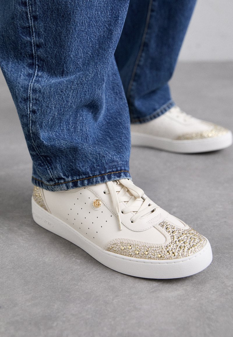 Weißer Sneaker mit strukturiertem Obermaterial, verziert mit goldenen Verzierungen an der Zehenpartie. Verfügt über Perforationen an den Seiten und eine weiße Sohle.
