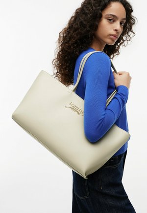 Donna con capelli lunghi e ricci che indossa un maglione blu e jeans scuri porta una grande borsa tote color crema Tommy Jeans sulla spalla.