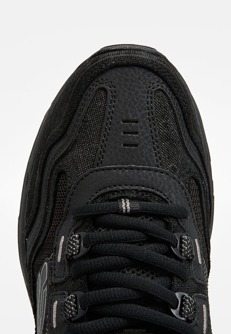 Chaussure de sport noire avec un dessus en mesh texturé, un embout renforcé et des lacets noirs proéminents. Présente des accents gris subtils et une semelle robuste.