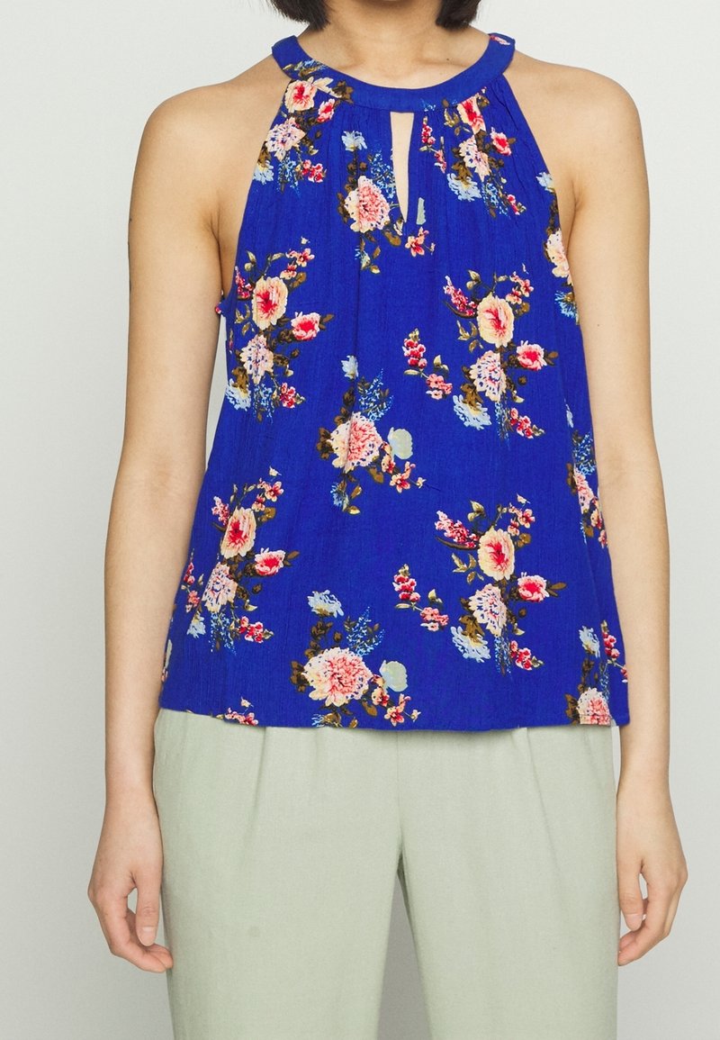 Modrý květinový halter top s klíčovou výřezem, s růžovými, bílými a zelenými květinami na texturované látce. V kombinaci se světlými kalhotami.