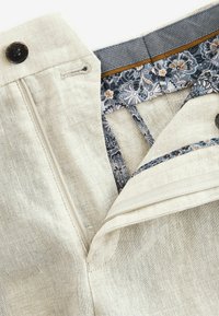 Pantaloni bej deschis cu un finisaj texturat din in, având o căptușeală cu model floral în albastru și alb și un sistem de închidere cu nasture închis la culoare.
