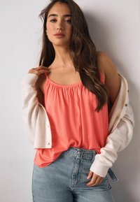 Koralų spalvos baklažanas su elastinga apykakle, dėvimas po baltu kardiganu. Šviesiai mėlyni džinsai su aukštu liemeniu, juostele juosmenyje ir laisvu siluetu.