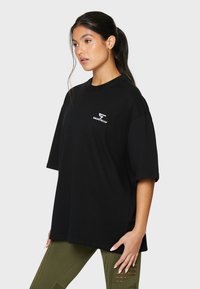 Sort oversized bomuld t-shirt med et hvidt logo på brystet, korte ærmer og en afslappet pasform. Enkel, glat tekstur.