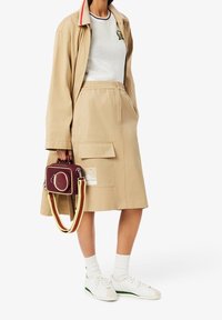 Trench-coat beige, haut en maille blanc, jupe beige avec poches, sac bordeaux avec accents jaunes, baskets blanches avec détail vert, chaussettes blanches.