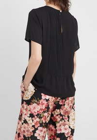 Blouse noire avec plis verticaux au dos, manches courtes et encolure ronde. Associée à un pantalon à motif floral.