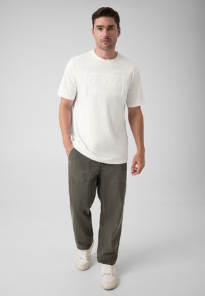 Homme portant une chemise blanche à manches courtes avec "GUESS" embossé sur la poitrine, un pantalon vert olive et des baskets blanches, debout sur un fond gris.