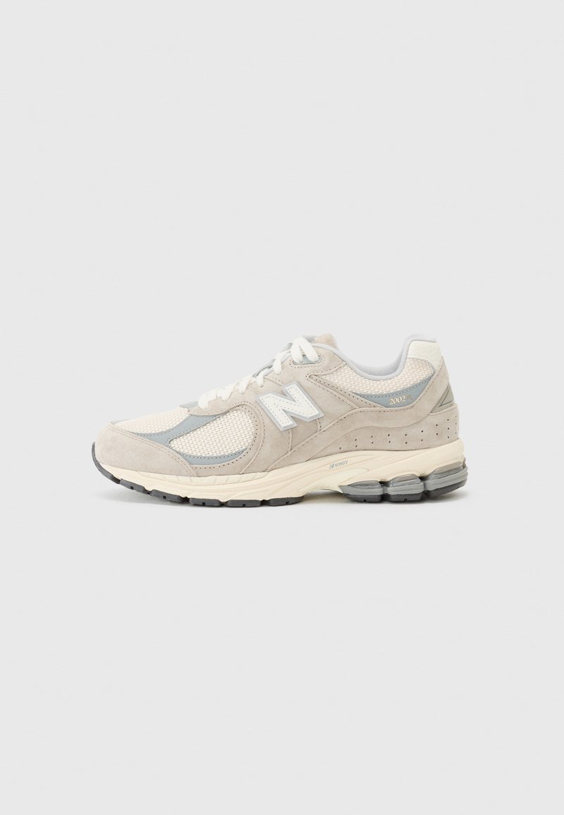 Chaussure de sport New Balance beige clair et grise avec des panneaux en mesh et en suède, lacets blancs et semelle rembourrée sur fond uni.