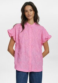 Blusa corta rosa a maniche corte con ricami floreali, maniche a volant svasate e chiusura con bottoni. Tessuto morbido e testurizzato visibile in tutto.