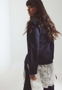 Giacca di jeans blu navy con maniche lunghe, dotata di un sovrapposto in pizzo bianco all'orlo. Il tessuto ha una consistenza liscia e una vestibilità strutturata.
