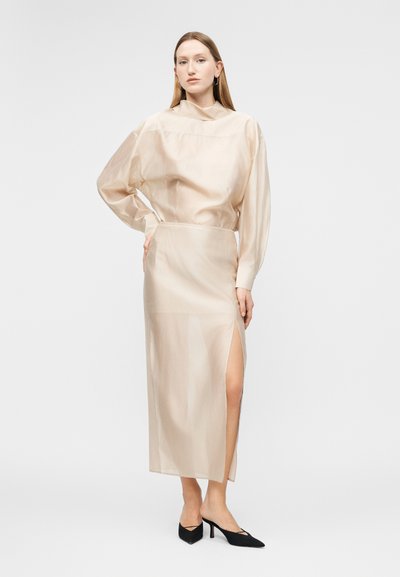 Femeie într-o rochie lungă bej transparentă, cu o crăpătură înaltă pe piciorul stâng, stând cu mâna pe șold, purtând tocuri negre ascuțite, pe un fundal simplu.