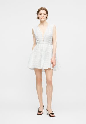 Γυναίκα που φοράει λευκό μίνι φόρεμα χωρίς μανίκια με υφή και βαθύ V λαιμόκοψη, μαζί με μαύρα ψηλοτάκουνα σανδάλια με λουράκια, στέκεται μπροστά σε μονόχρωμο φόντο.