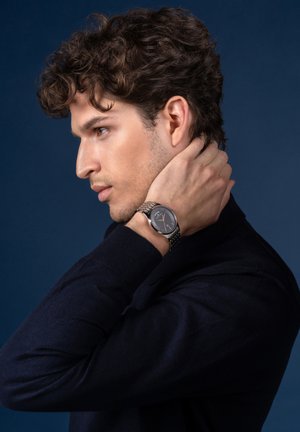 Silberne Uhr mit rundem Ziffernblatt, schwarzem Ziffernblatt, strukturiertem Muster und Metallarmband, das am Handgelenk getragen wird. Dunkler Pullover vor blauem Hintergrund.