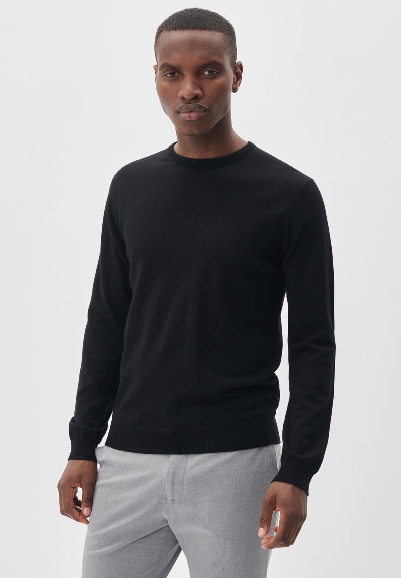 Melnā adīta džempera ar apaļo kakla izmēru un garām piedurknēm, kuram ir gluda tekstūra. Apvienots ar gaiši pelēkām biksēm.