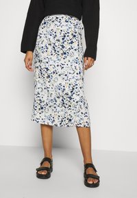 Jupe midi florale blanche, ornée de fleurs bleues et jaunes, avec un léger reflet et un détails de nœud sur le côté. Associée à des sandales noires.