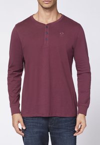 Bordeaux rotes Langarm-Henley-Shirt aus weicher Baumwolle mit drei Knöpfen am Hals und einem gestickten Logo auf der Brust. Getragen mit Jeans.