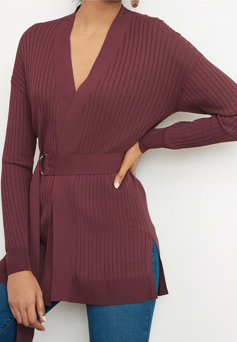 Cardigan côtelé bordeaux avec un décolleté en V profond, des manches longues et une ceinture nouée à la taille. Présente des fentes latérales et une texture douce.