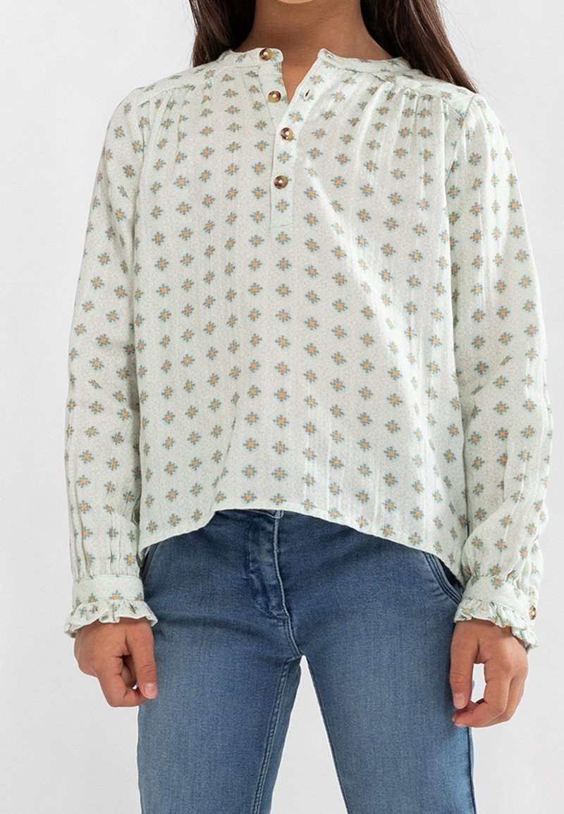 Femme portant un chemisier à fleurs vert clair avec des boutons marron et des poignets à volants, associé à un jean en denim bleu.