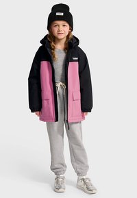 Färgblockerad jacka i svart och rosa, med dragkedja, framfickor och huva. Bärs över en grå outfit med en svart mössa.