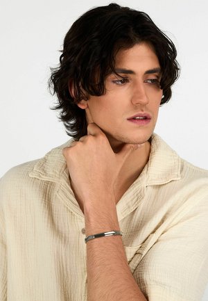 Bracciale in metallo color argento con un design elegante e piatto, arricchito da sottili accenti geometrici. Indossato su un polso contro una camicia beige testurizzata.