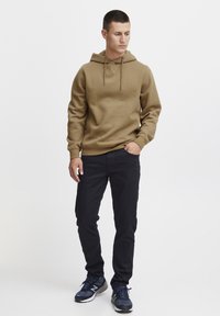 Brown hoodie with a drawstring, paired with black pants and blue sneakers. The hoodie features a textured material and a relaxed fit.
