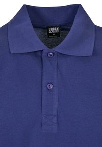 Polo en coton violet avec un col, comportant deux boutons au niveau du plastron et un tissu texturé. Étiquette de la marque visible sur le col.