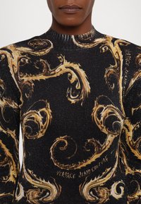Maglione nero con un elaborato motivo a spirale dorato, dotato di collo a girocollo a costine e texture scintillante. È visibile il marchio "Versace Jeans Couture".