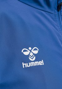 Blå sportjacka med en slät textur och en framträdande vit logotyp med en stiliserad design på vänster bröst.