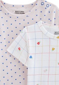 Deux t-shirts empilés : l'un est rose pâle avec de petits cœurs bleus, l'autre est blanc avec un motif à carreaux coloré et des accents floraux. Matériau en coton doux.
