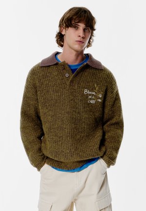 Olivgrüner, gestrickter Pullover mit braunem Kragen, drei Knöpfen und dem gestickten Schriftzug "Blooming with Care" auf der Vorderseite.