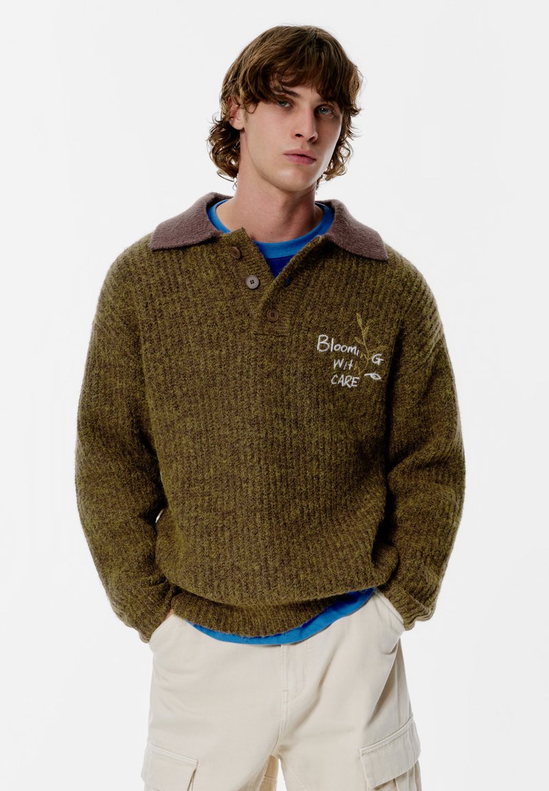 Oliivinvihreä neulottu neule, jossa on ruskea kaulus, kolme nappia ja etuosassa kirjailtu "Blooming with Care" -teksti.