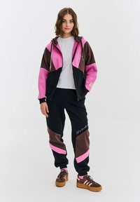 Młoda kobieta stoi, ubrana w różową, brązową i czarną bluzę z kolorowym blokiem oraz pasujące dresy z brązowymi sneakersami i różowymi skarpetkami.