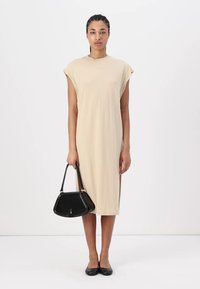 Robe en coton beige avec des manches courtes et raglan, coupe à mi-genoux, portée avec un sac à main noir et des ballerines noires. Texture lisse, design simple.