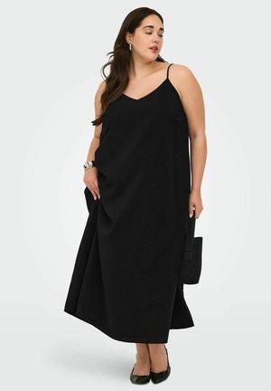 Frau trägt ein langes schwarzes ärmelloses Kleid mit dünnen Trägern, hält eine schwarze Handtasche und trägt schwarze spitze Schuhe.