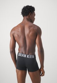 Melnas kokvilnas bokseris ar balto "CALVIN KLEIN" logotipu uz jostas, izcelti uz kailas muguras pret vienkāršu fona.