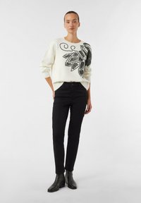 Kremowy sweter z czarnym liściastym wzorem, w połączeniu z czarnymi obcisłymi dżinsami i czarnymi botkami za kostkę. Sweter ma okrągły dekolt i ściągacze przy mankietach.