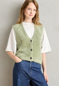 Getextureerde groene vest met v-hals, vijf zwarte knopen, gedragen over een wit shirt. Blauwe jeans maken de outfit compleet. Effen achtergrond.