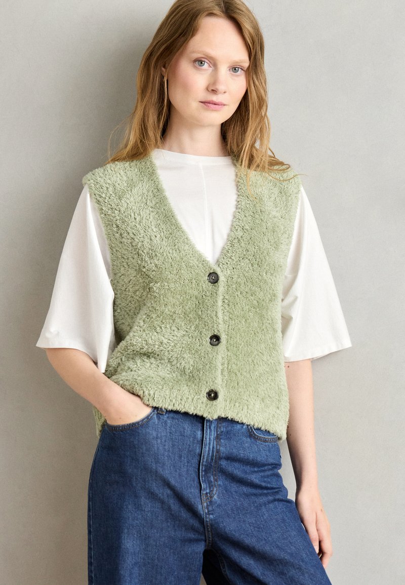 Getextureerde groene vest met v-hals, vijf zwarte knopen, gedragen over een wit shirt. Blauwe jeans maken de outfit compleet. Effen achtergrond.