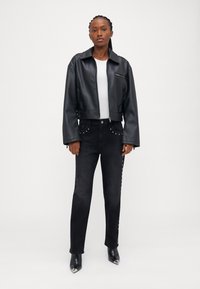 Jachetă cropped din piele sintetică neagră, cu guler, purtată cu blugi negri cu studuri decorative. Modelul poartă cizme negre cu vârf ascuțit.
