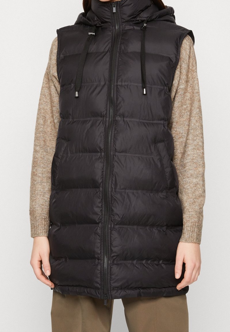 Svart pufferväst med huva, kviltat mönster, framdragkedja och justerbara snören. Bärs över en ljusbrun texturerad tröja.