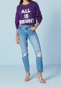 Pull violet avec l'inscription "ALL IS BRIGHT" en sequins colorés, associé à un jean bleu clair déchiré et des sandales à talons transparents.