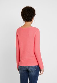 Różowy sweter o prążkowanej fakturze z długimi rękawami i szerokim dekoltem, w zestawieniu z niebieskimi dżinsami. Materiał wydaje się miękki, z delikatną teksturą.