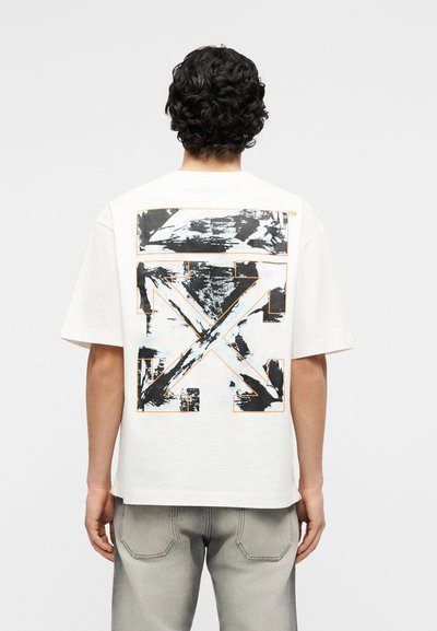 Άνδρας που φοράει λευκό υπερμεγέθες T-shirt με μαύρο και πορτοκαλί αφηρημένο βέλος στην πλάτη, σε συνδυασμό με ανοιχτόγκρι τζιν.